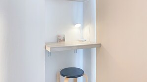 Een hoek van een kamer met een witte muur, een houten plank met een fotolijstje en een lamp, en een houten kruk.