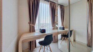 Une chambre d’hôtel moderne avec un bureau en bois, deux chaises noires, un grand miroir et une vue sur la ville à travers des rideaux transparents.