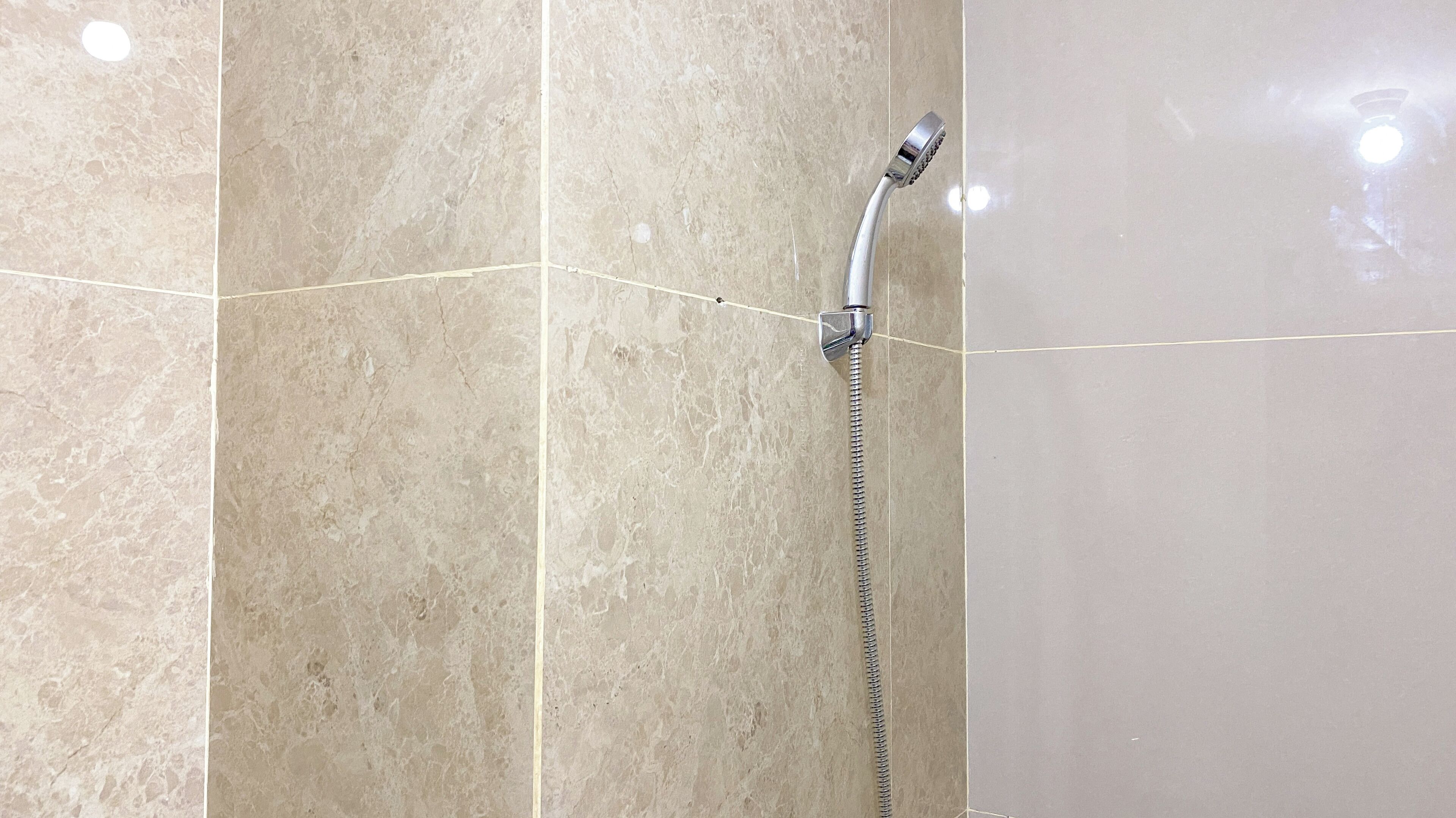 A shower with a chrome head and a flexible hose, mounted on a wall with beige marble tiles.