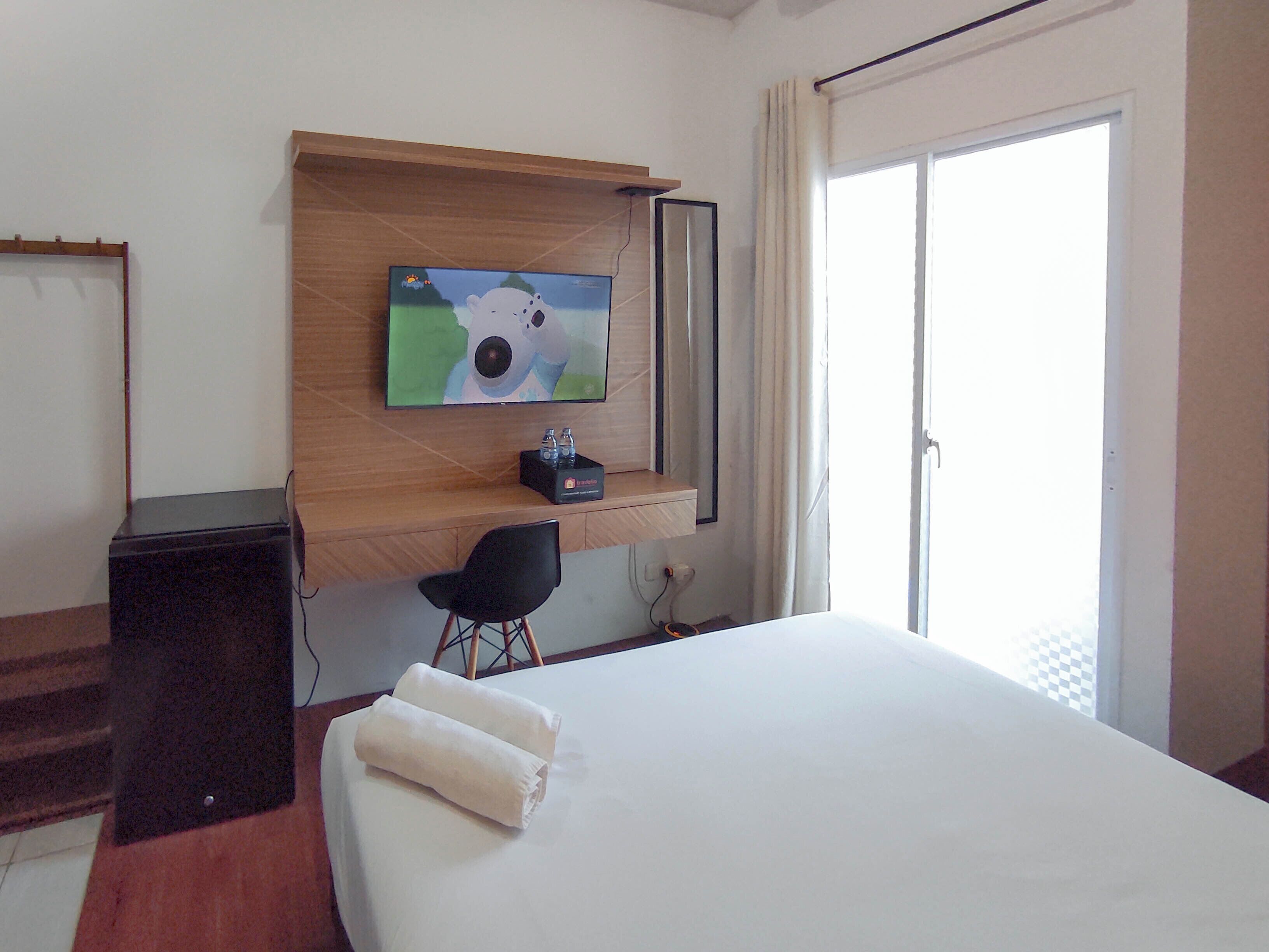 A hotel room with a bed, a desk, a chair, a TV mounted on a wooden shelf, and a window with curtains.