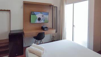 A hotel room with a bed, a desk, a chair, a TV mounted on a wooden shelf, and a window with curtains.