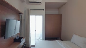 A hotel room with a bed, a TV mounted on the wall, a wardrobe, and a sliding glass door leading to a balcony. - Homey Studio With Easy Pool Access Apartment At Suncity Residence (Sidoarjo)