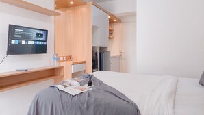 A hotel room with a bed, a TV mounted on the wall, a wooden shelf with a water bottle, and a small table with a magazine and a candle. - Good Place Studio At Urbantown Serpong Apartment (South Tangerang)