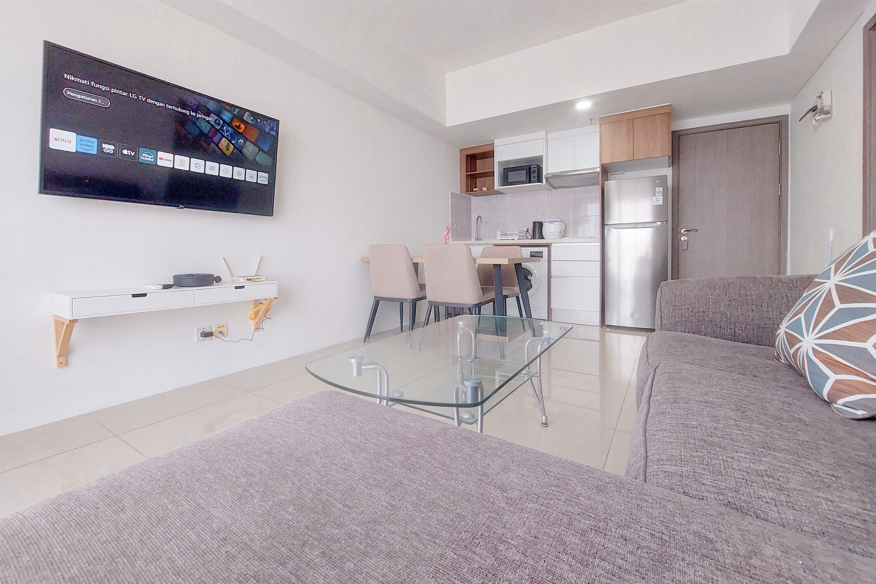 A modern living room with a glass-top coffee table, a flat-screen TV mounted on the wall, a sofa, and a dining area with chairs.