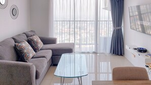 A modern living room with a grey sofa, patterned cushions, a glass coffee table, and a view through large windows. - Comfortable And Homey 1Br Tamansari Bintaro Mansion Apartment (South Tangerang)