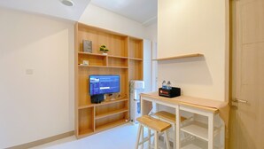 A modern room with a wooden shelving unit, a small desk with a stool, and a television displaying news. - Cozy Living 2Br Apartment At Tokyo Riverside Pik 2 (Teluknaga)