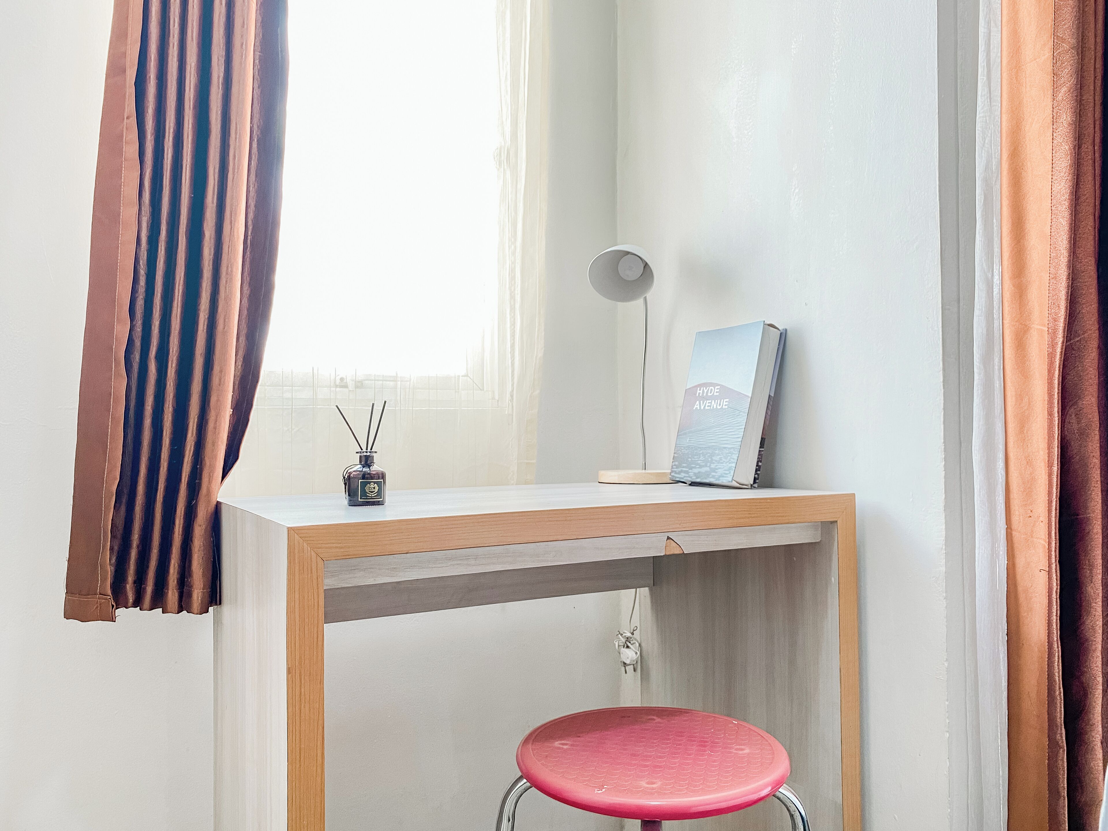 Ein kleiner Arbeitsbereich mit einem Holztisch, einem roten Hocker, einer Lampe, einem Buch mit dem Titel 'HYDE AVENUE' und einer Rattan-Diffusorflasche.