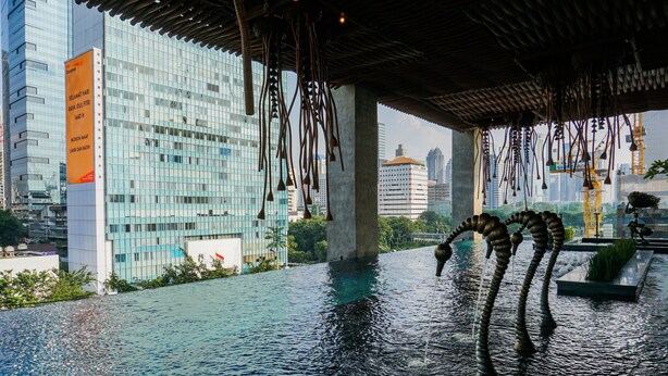 Un bâtiment moderne doté d’une façade en verre réfléchissant, d’un élément décoratif aquatique agrémenté d’une sculpture et offrant une vue sur le paysage urbain.