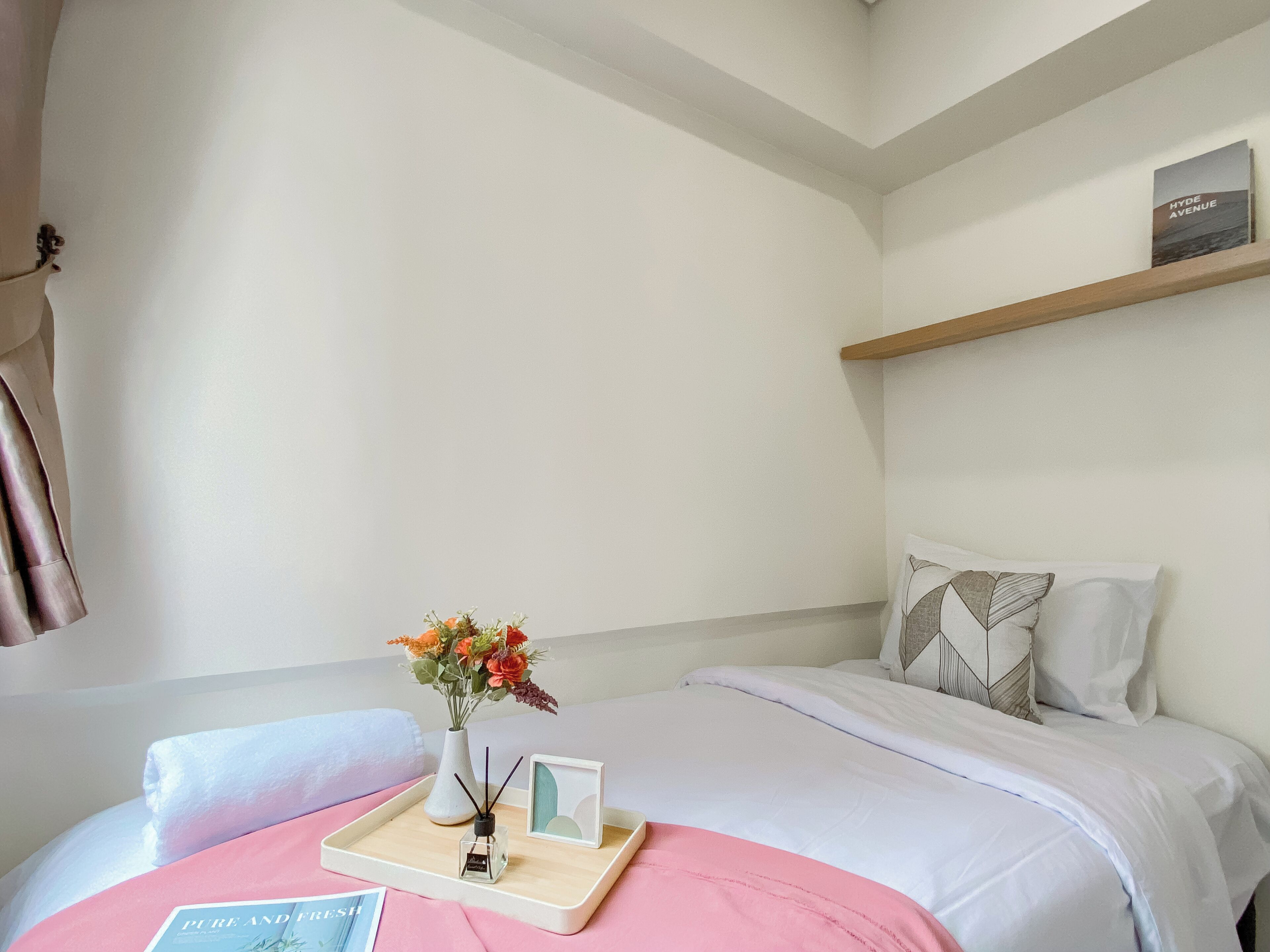 Ein Hotelzimmer mit einem Bett, einem Nachttisch mit Blumenvase, einem Tablett mit Buch und Kerze sowie einem Regal mit dem Buch ‚Hyde Avenue‘.