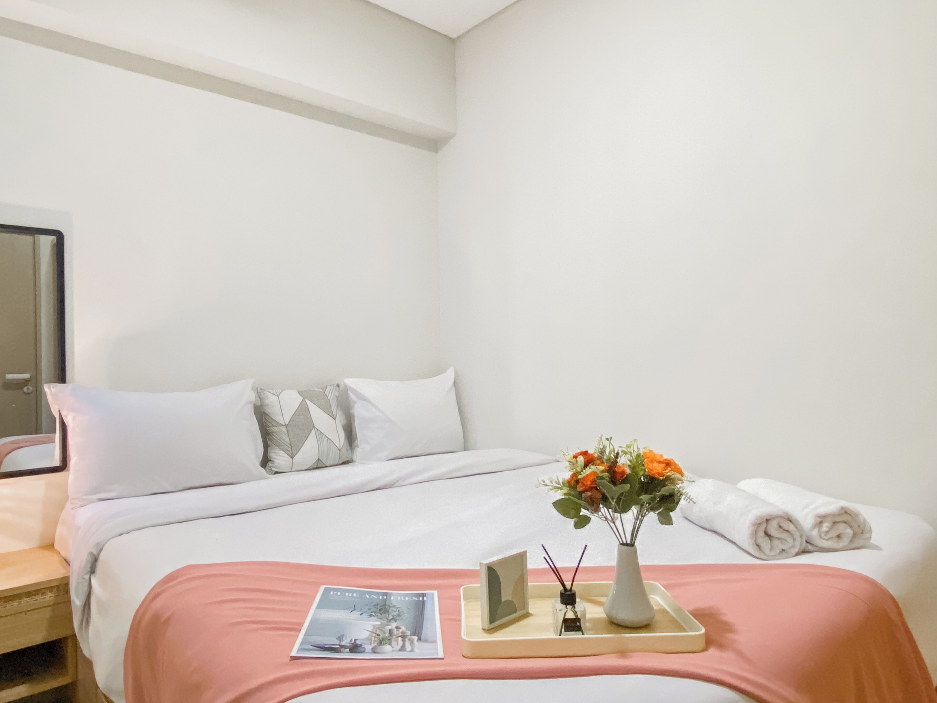 A neatly made bed with white and gray pillows, a pink bedspread, a small vase with flowers, and a rolled towel on the bed.