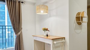 A small, modern room with a wooden desk, a white chair, a hanging light fixture, and a view of a building through a window with curtains. - Well Furnished And Comfort 2Br Meikarta Apartment (Cikarang)