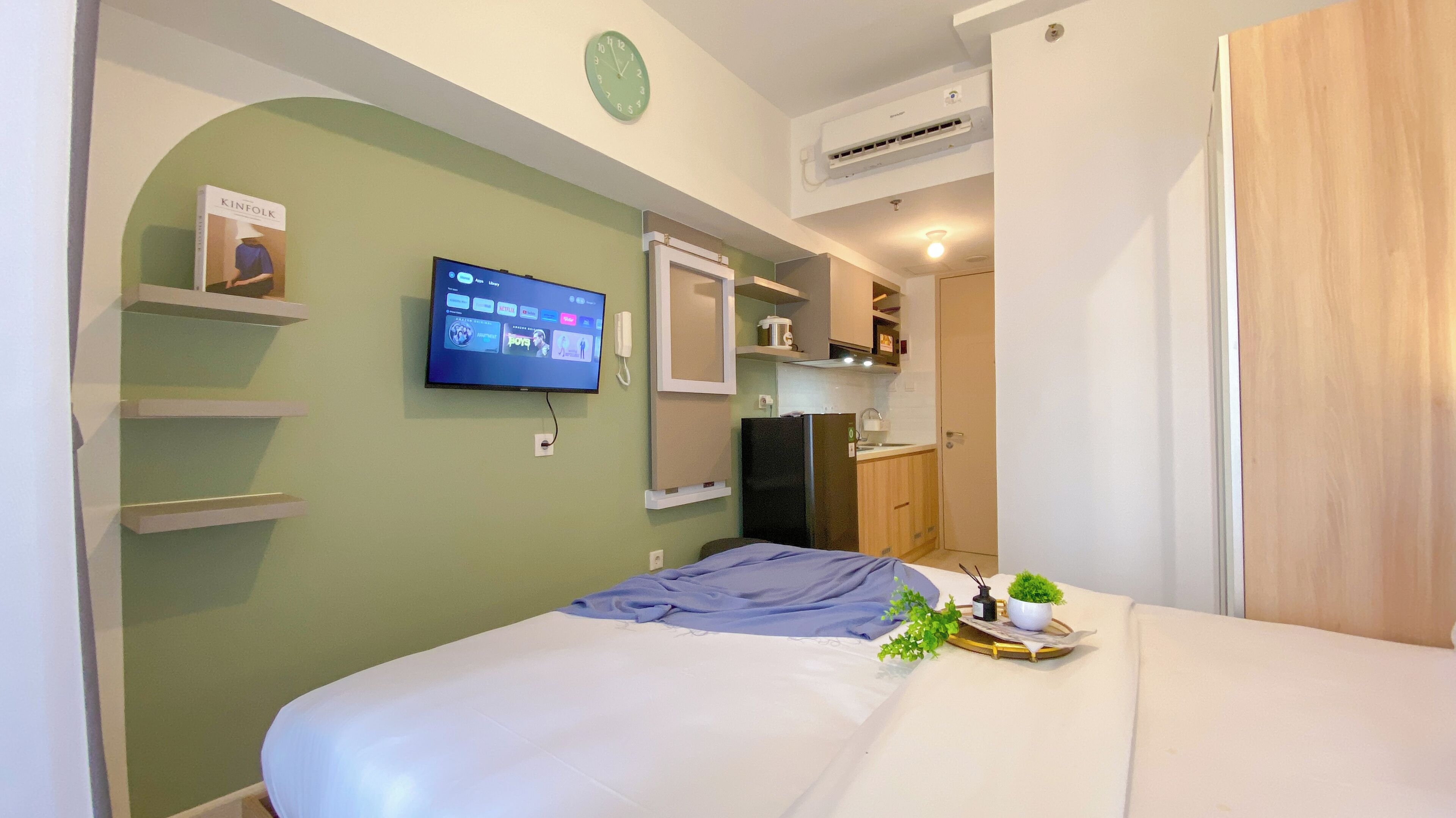 A bedroom with a bed, a TV mounted on the wall, a small kitchen area, and a door leading to another room.