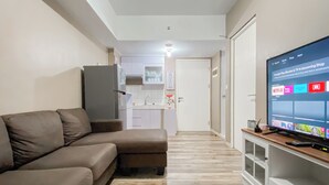Un salon avec un canapé marron, un téléviseur posé sur un meuble et une cuisine à l’arrière-plan.