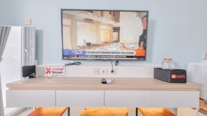 Une chambre d’hôtel moderne équipée d’un téléviseur à écran plat fixé au mur, diffusant une chaîne d’information, d’un bureau en bois avec une télécommande, d’une valise de voyage et de deux tabourets en bois.