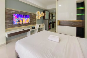 A modern hotel room with a large bed, a TV mounted on the wall, a desk with a chair, and a kitchenette area. - The Modern Studio Room At Beverly Dago Apartment (Bandung)