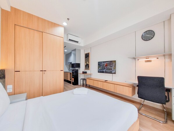 A hotel room with a bed, a desk, a chair, a TV, a clock, and a kitchenette in the background. - Good And Comfort Stay Studio Patraland Amarta Apartment (Ngaglik)