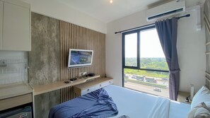 A modern hotel room with a bed, a TV mounted on a wooden panel wall, a desk with a computer, and a large window with a view of a highway. - Homey And Clean Studio At Sky House Alam Sutera Apartment (Tangerang)