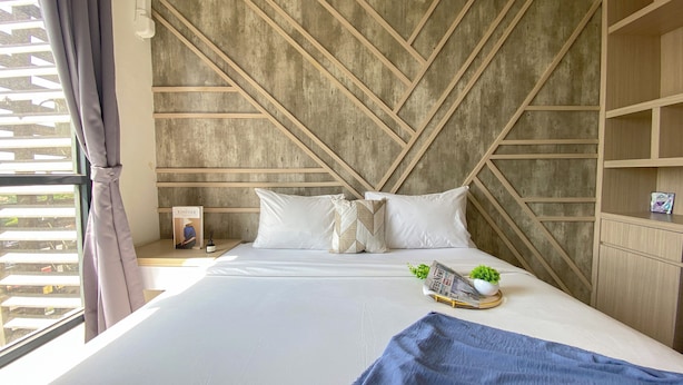 A modern bedroom with a geometric wooden headboard, a neatly made bed with white linens, a small bedside table with a book and a candle, and a window with curtains. - Homey And Clean Studio At Sky House Alam Sutera Apartment (Tangerang)