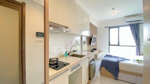 A compact kitchen with a stove, sink, and microwave. There is a window with curtains and a bed with a blue blanket. - Homey And Clean Studio At Sky House Alam Sutera Apartment (Tangerang)