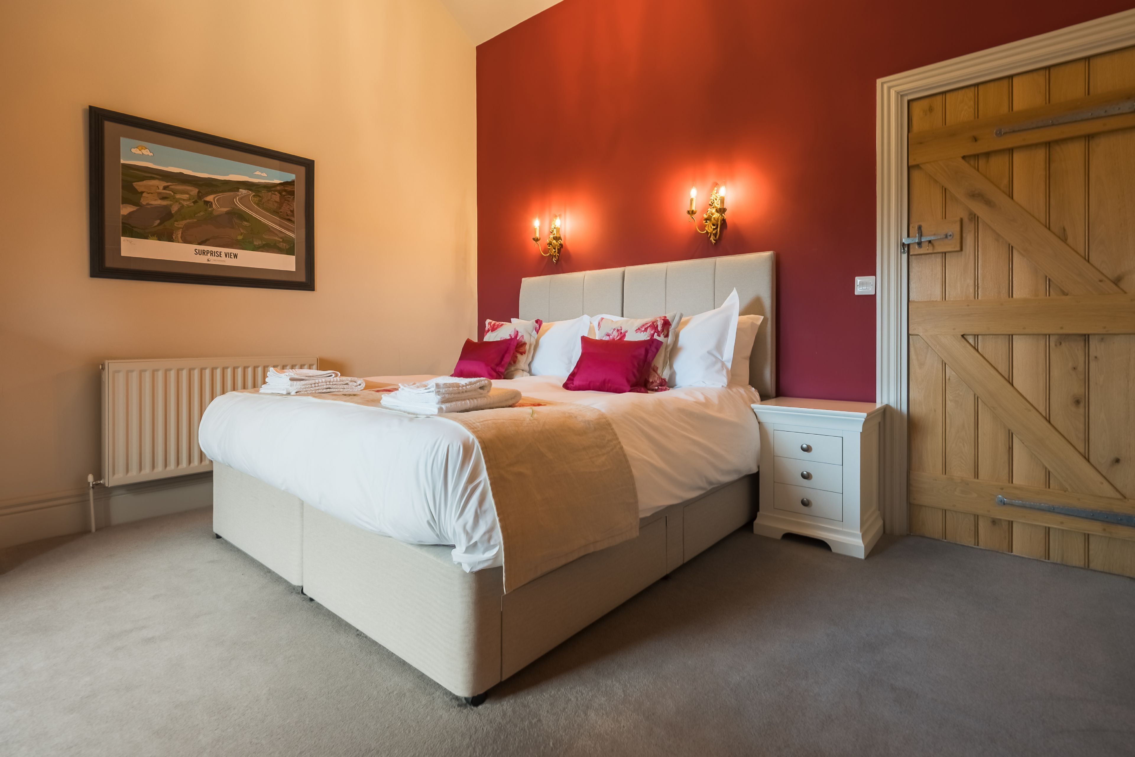 A bedroom with a large bed, a bedside table, a nightstand, a framed picture on the wall, and a wooden door.