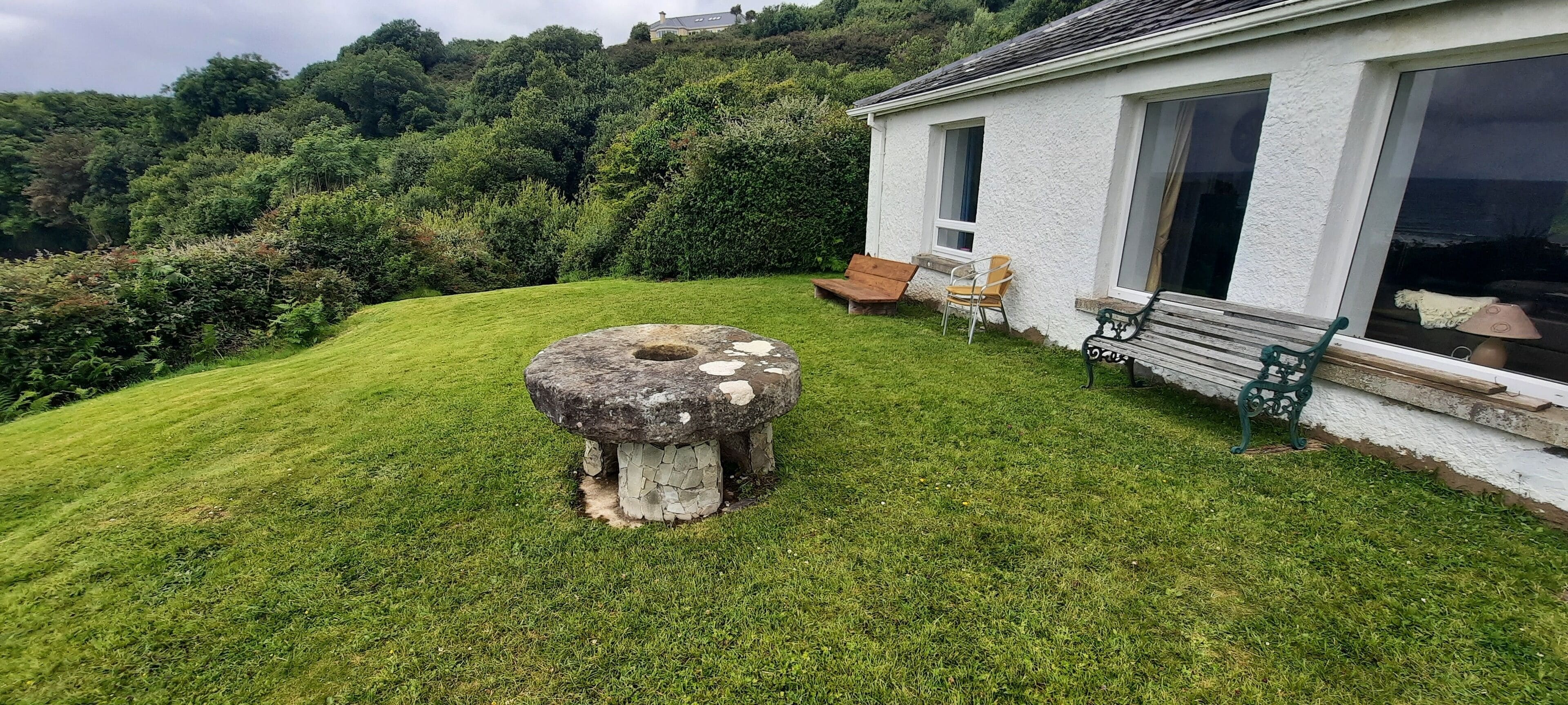 Ein steinerner Brunnen in einer grünen Wiese, im Hintergrund ein weißes Gebäude.