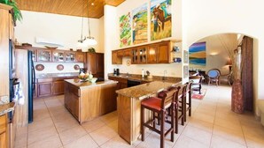 A spacious kitchen with wooden cabinets, a granite countertop island, and a dining area with chairs. - Luxury Villa | Infinity Pool & Ocean Views (Playa Potrero)
