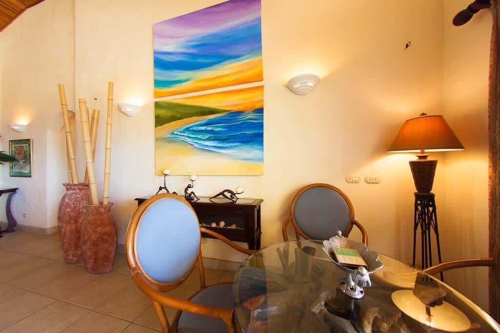 Un coin repas comprenant une table en verre, deux chaises, un tableau représentant un paysage et une lampe.