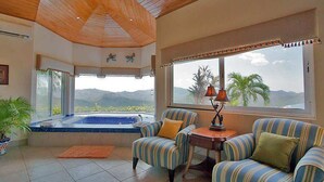 A room with a wooden ceiling, a large window with a view of palm trees and mountains, a hot tub, and striped armchairs. - Luxury Villa | Infinity Pool & Ocean Views (Playa Potrero)