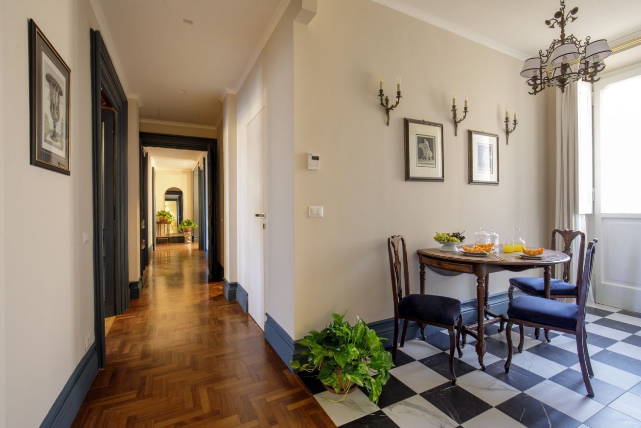 A hallway with wooden flooring leading to a dining area with a checkered floor, a table with chairs, and wall-mounted lighting.