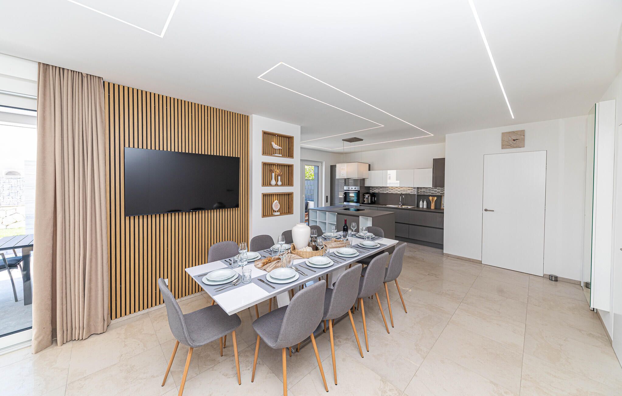 A modern dining area with a large table set for six, a flat-screen TV mounted on a wooden panel wall, and a kitchen in the background.