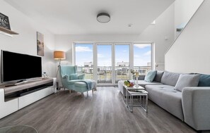 Een moderne woonkamer met een flatscreen-tv, een lichtblauwe fauteuil, een grijze bank en een kleine tafel met een fruitschaal.