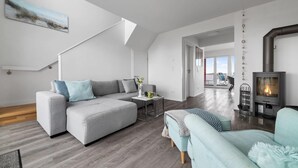 Un salon moderne avec un canapé gris, un fauteuil bleu clair et un plancher en bois. Un escalier mène à un balcon, et un poêle à bois se trouve dans un coin.