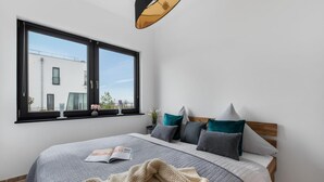 Une chambre à coucher avec un grand lit, une fenêtre donnant sur un immeuble, et une table de chevet avec une lampe et des fleurs.
