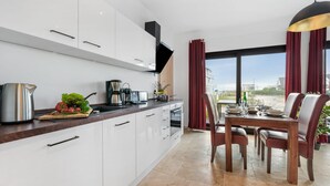 Une cuisine moderne avec des meubles blancs, un coin repas avec une table dressée pour quatre personnes, et une grande fenêtre donnant sur des immeubles.