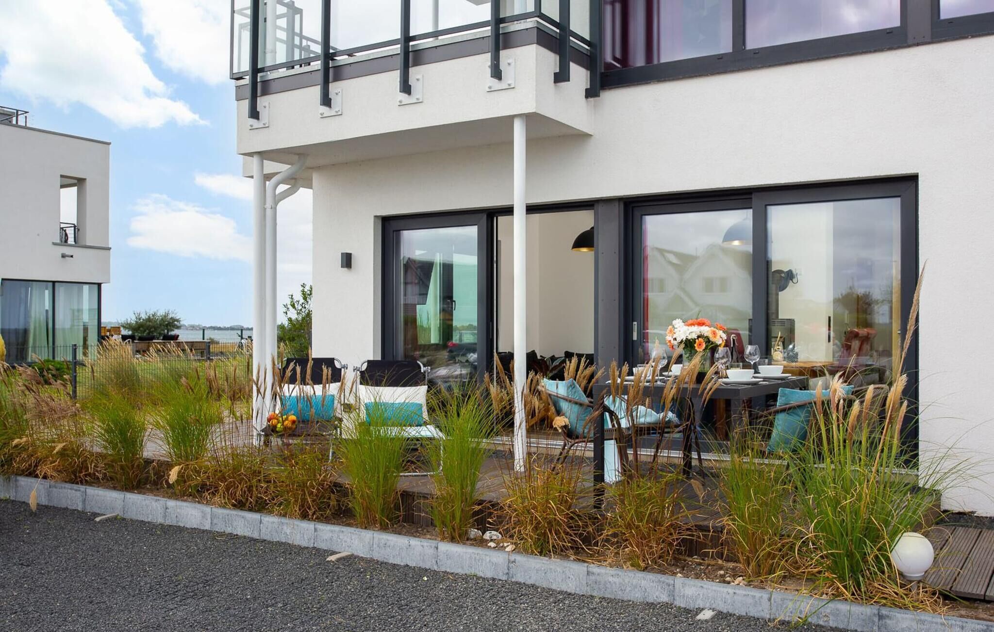 A modern outdoor dining area with a table set for two, surrounded by tall grasses and a concrete border.