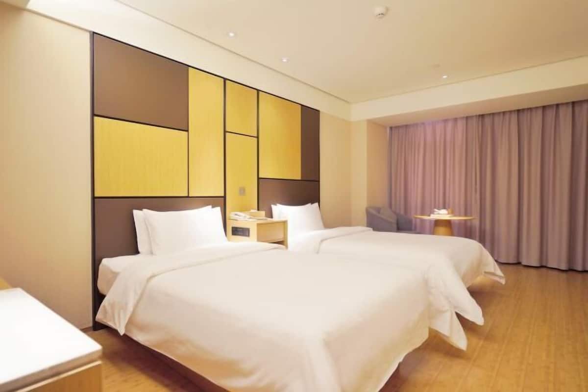 A hotel room with two beds, a large window with curtains, a bedside table with a lamp, and a small table with a lamp.