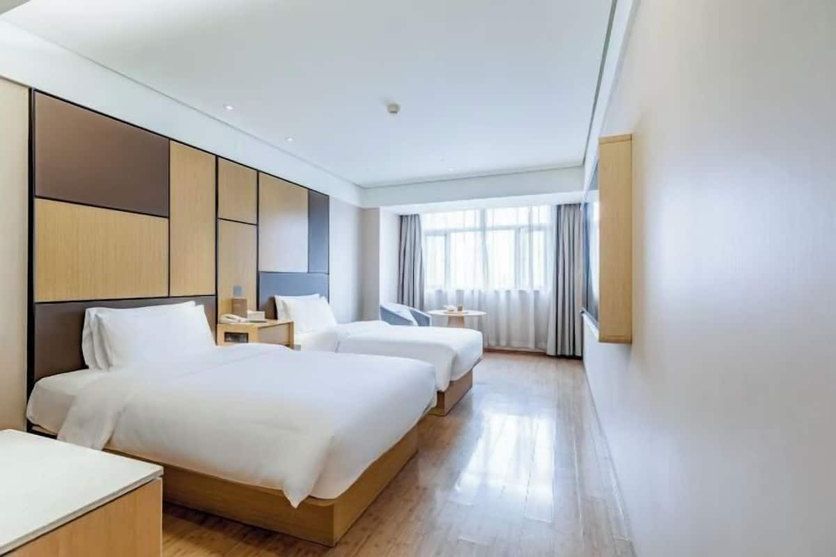 A hotel room with two beds, a bedside table with a lamp, a small round table, and a window with curtains.