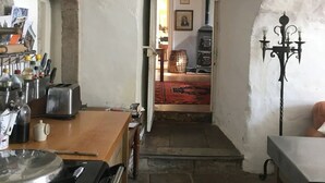 Une cuisine rustique avec un sol en pierre, une table en bois et un poêle. Une porte mène à un salon agrémenté d’un tapis et d’une lampe.