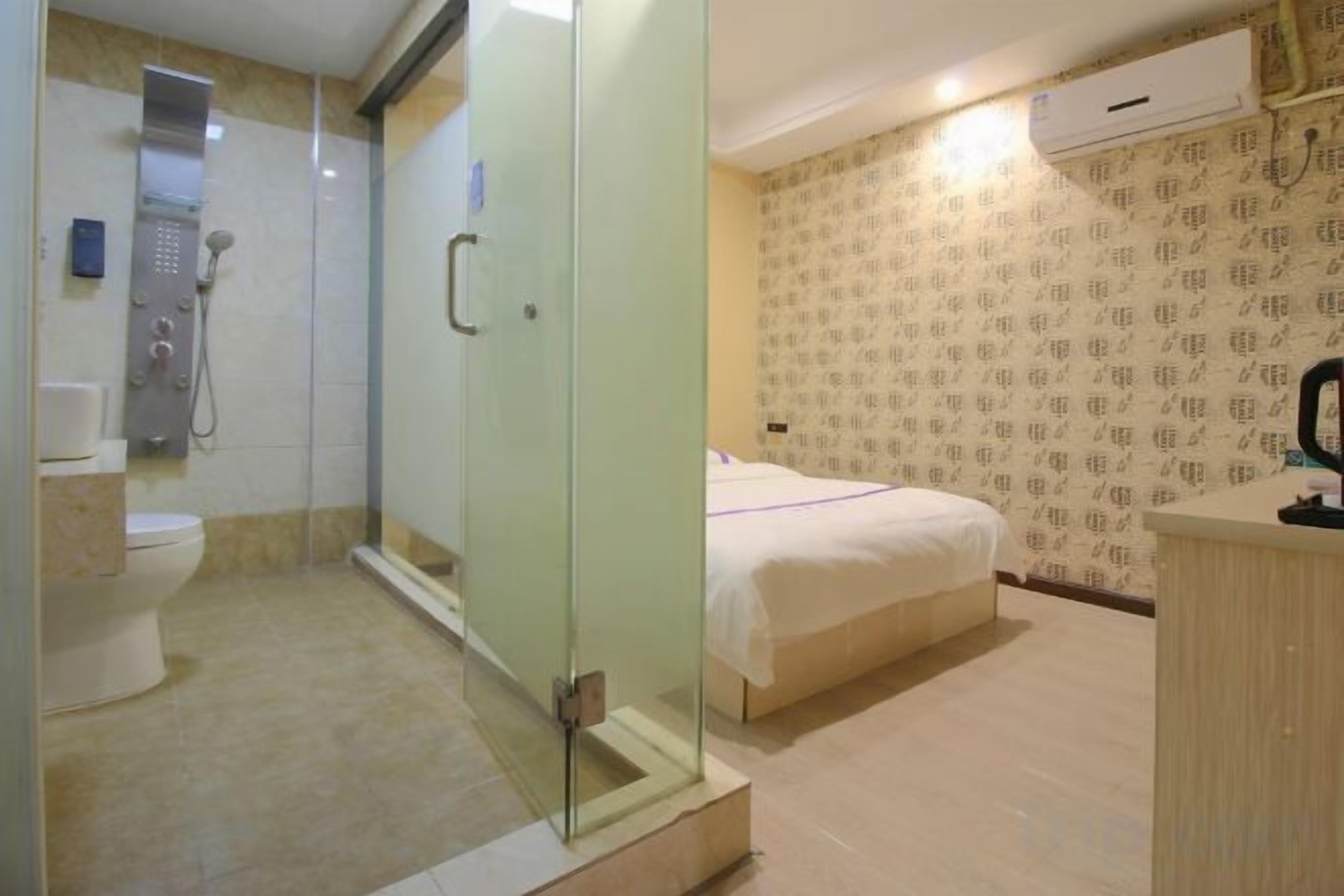 A bathroom with a glass-enclosed shower, a toilet, and a bed with a white bedspread.