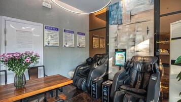 Une salle de massage équipée de deux fauteuils de massage, d’une table en bois ornée d’un vase de fleurs et de panneaux d’information accrochés au mur.
