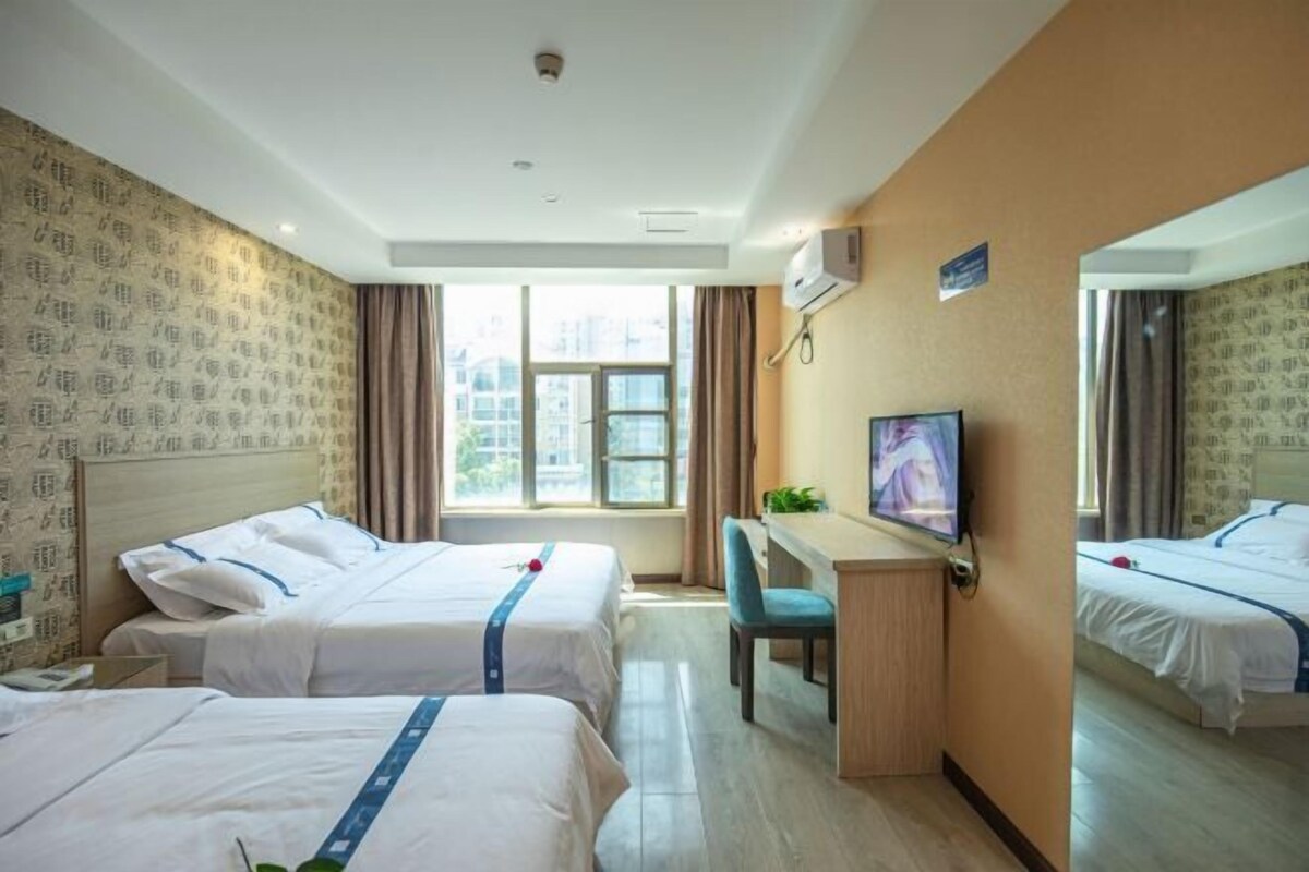 A hotel room with two beds, a desk, a TV, and a window with a view of buildings.