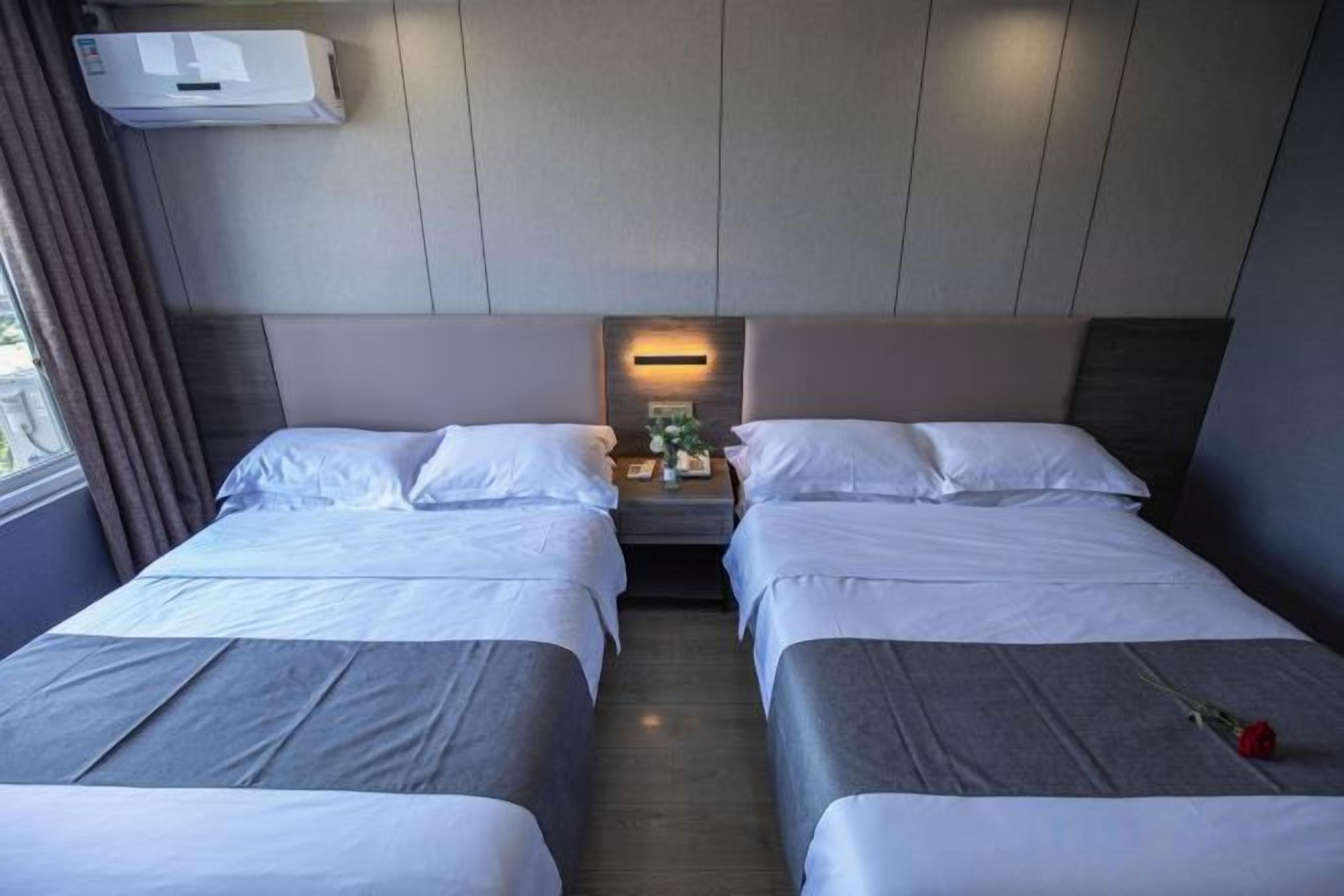 A hotel room with two beds, a bedside table with a lamp, and a window with curtains.