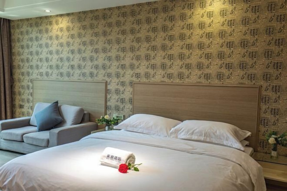 A hotel room with a large bed, two pillows, a bedside table, a lamp, and a floral arrangement.