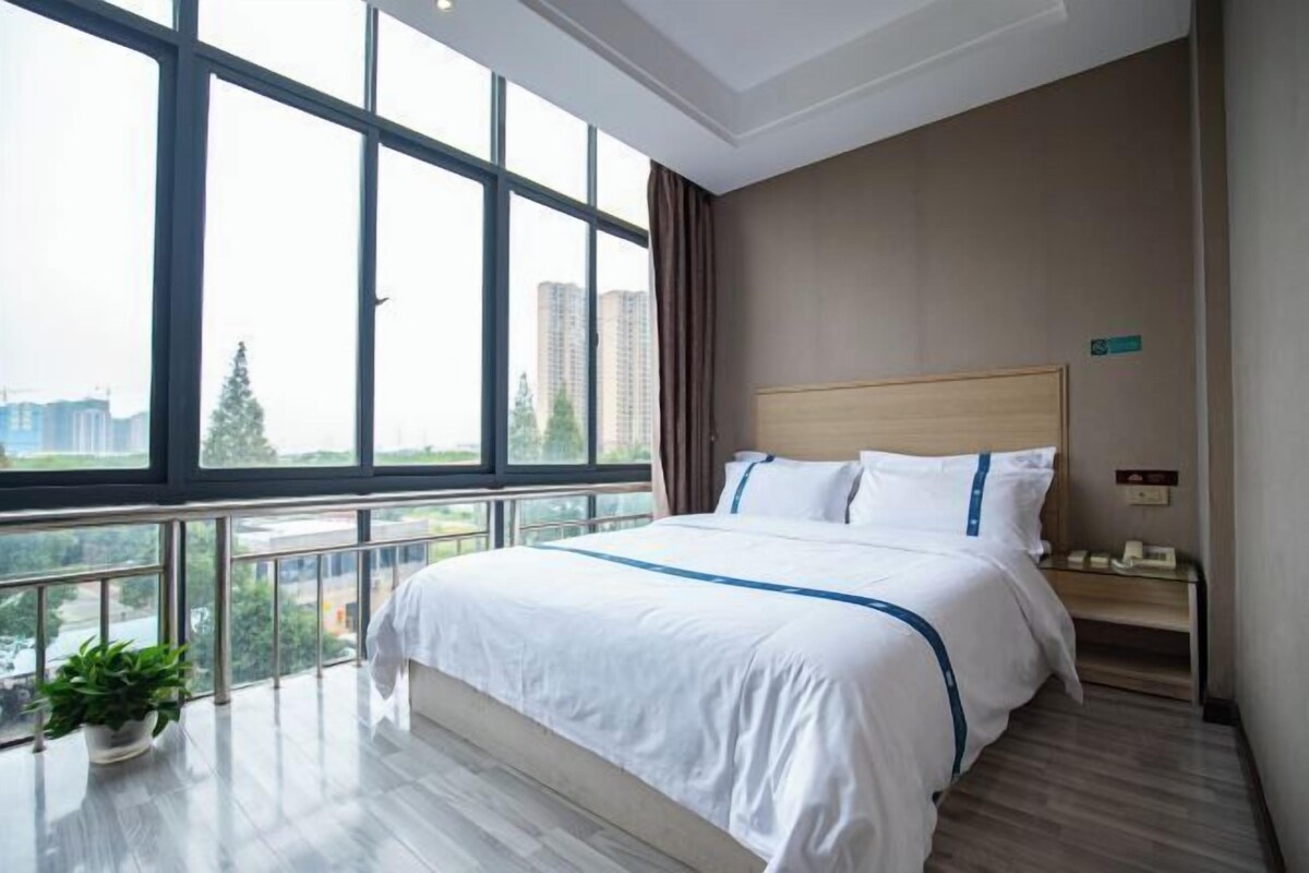 A hotel room with a large window, a bed with white bedding, a wooden headboard, a bedside table, and a potted plant.