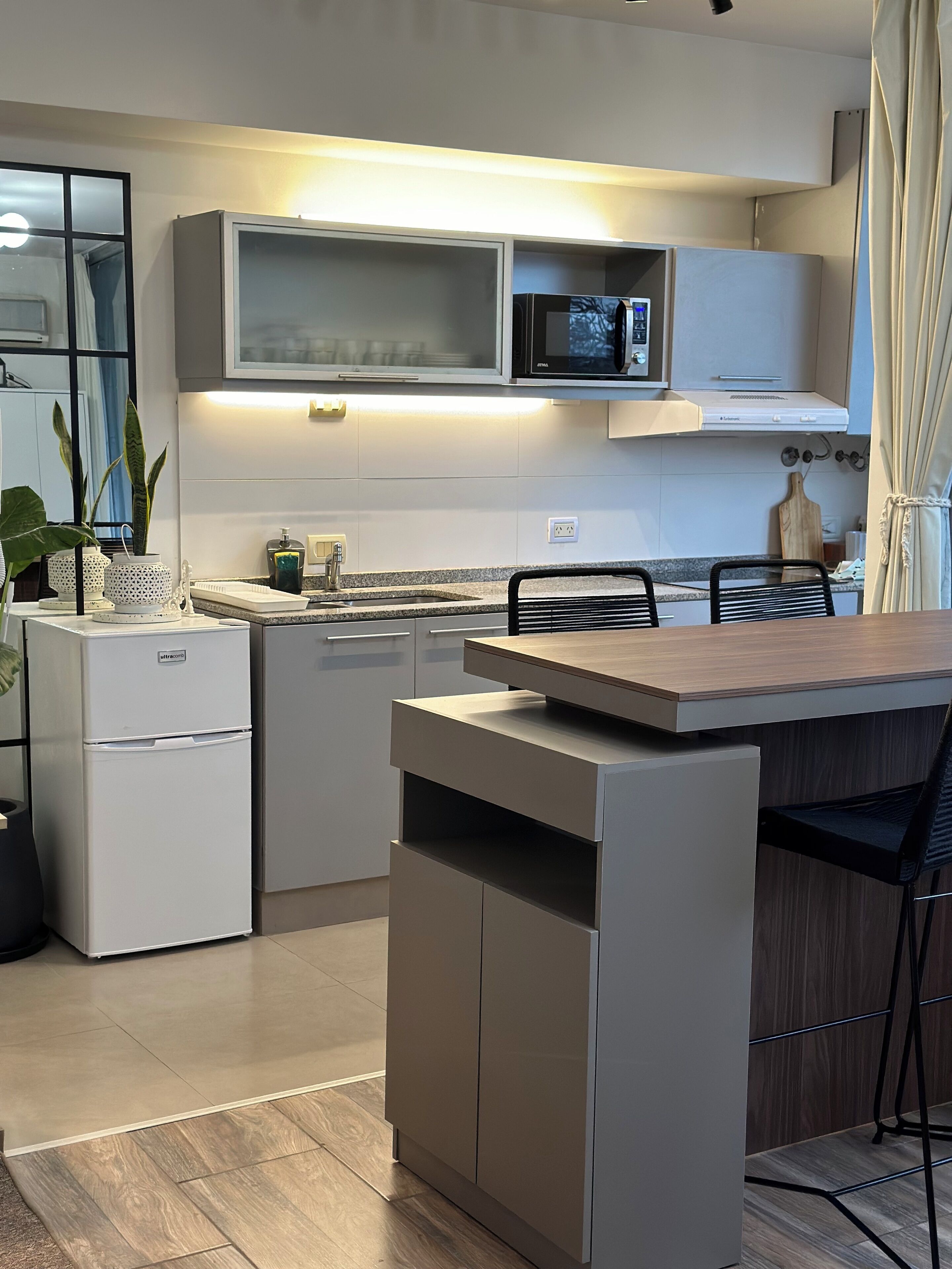 Una cocina compacta con microondas integrado, refrigerador y una mesa de comedor de madera con sillas.