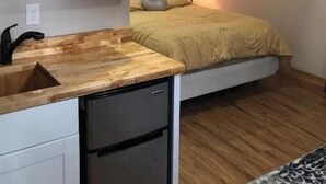 Une chambre à coucher de taille réduite, comprenant un lit, une lampe de chevet et une petite kitchenette  équipée d’un évier et d’un réfrigérateur.