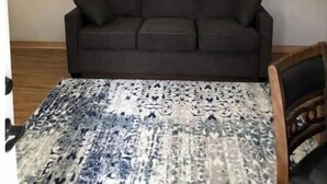 Une pièce avec un canapé marron foncé placé contre un mur uni, un tapis à motifs et une poutre en bois au plafond.