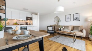 A modern kitchen with white cabinets, a dining table with chairs, a sofa, and a coffee table. There are framed pictures on the wall and a potted plant on the floor.