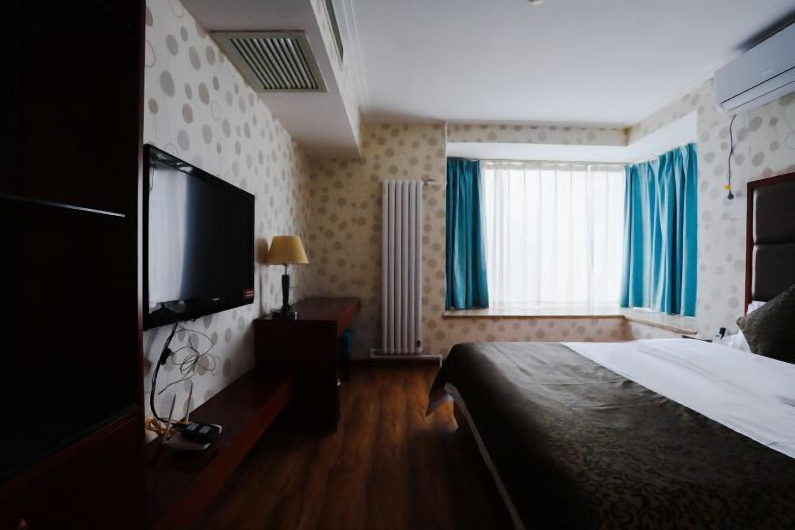 A hotel room with a bed, a television, a window with curtains, and a radiator.