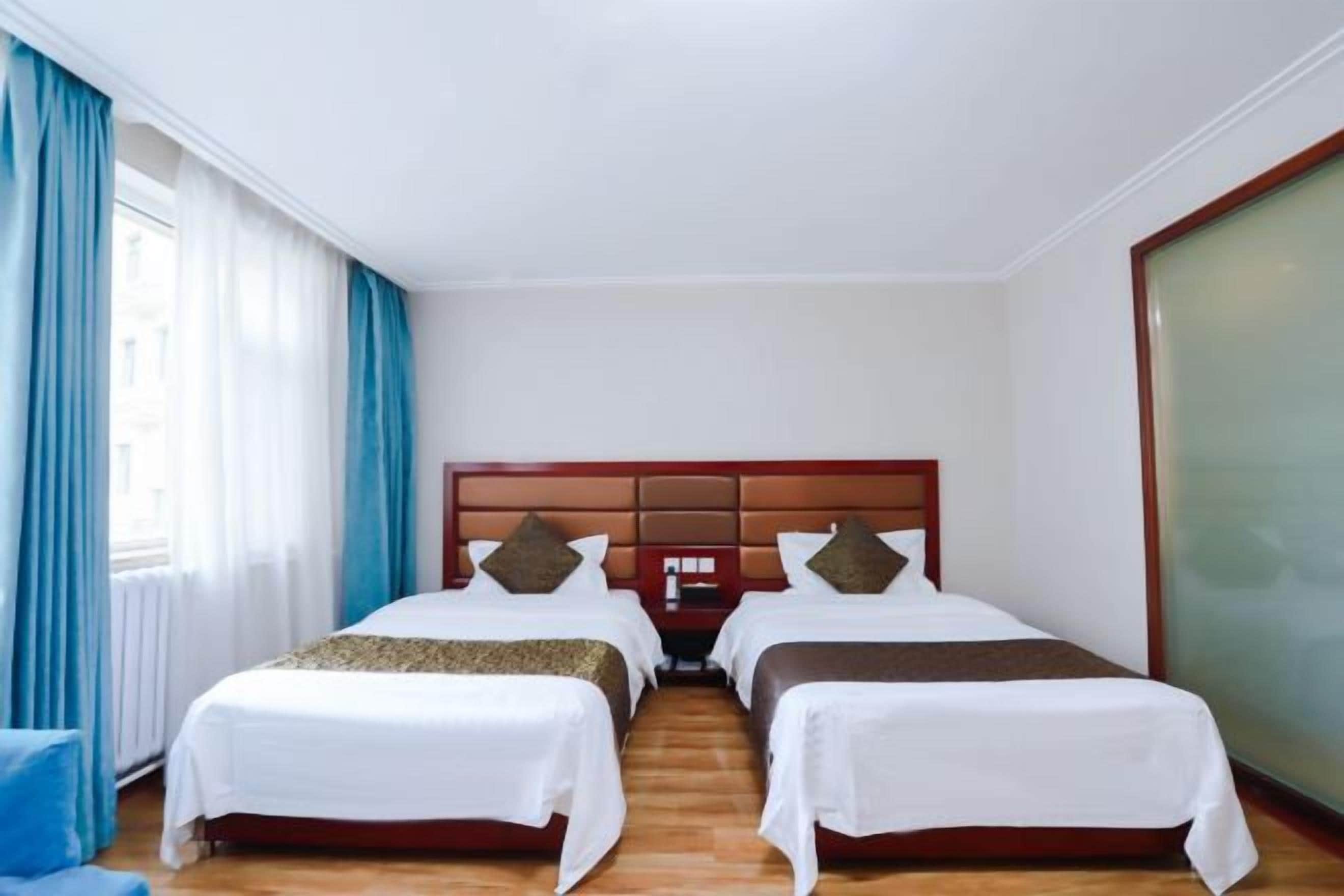A hotel room with two single beds, a wooden headboard, a large window with curtains, and a view of the city.
